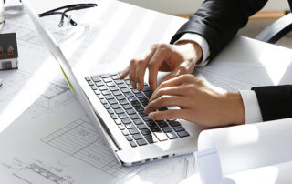 Hands of CEO of construction company in black suit typing message via e-mail to partners on laptop with drawings, seal stamp, divider on table. Young architect using netbook sitting in his office